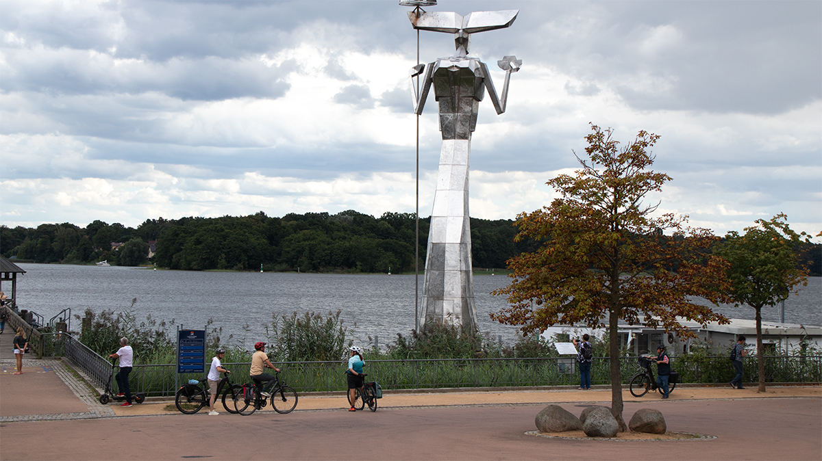 Edelstahlskulptur Parzival am Ruppiner See mit Baum, Uferbereich und dunklen Wolken im Hintergrund.